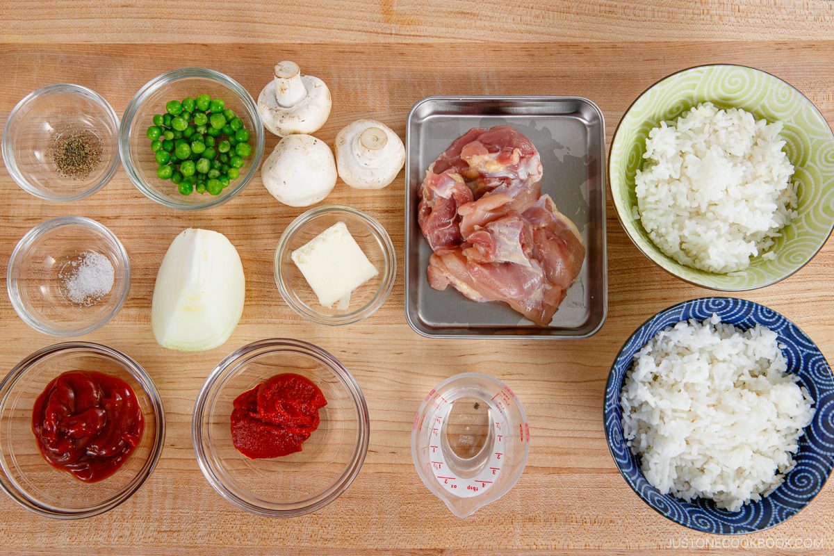 Various ingredients on a wooden surface—raw chicken, two bowls of white rice, mushrooms, peas, onion, butter, ketchup, tomato paste, black pepper, and a measuring cup with liquid—are gathered together as the essential components for making Japanese Chicken Rice (Midnight Diner).