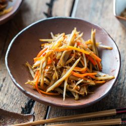 A bowl of Kinpira Gobo (Braised Burdock Root), a classic Japanese dish featuring sautéed burdock root and carrots, sits on a wooden table with chopsticks beside it.