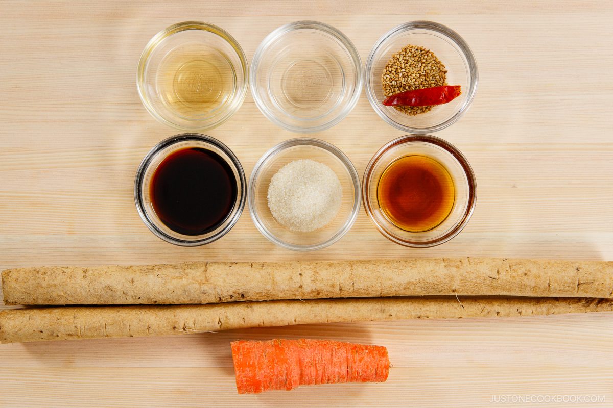 Six small bowls with sauces, vinegar, sugar, and sesame seeds are arranged above two long burdock roots and a piece of carrot on a light wooden surface—perfect ingredients for preparing Kinpira Gobo (Braised Burdock Root).
