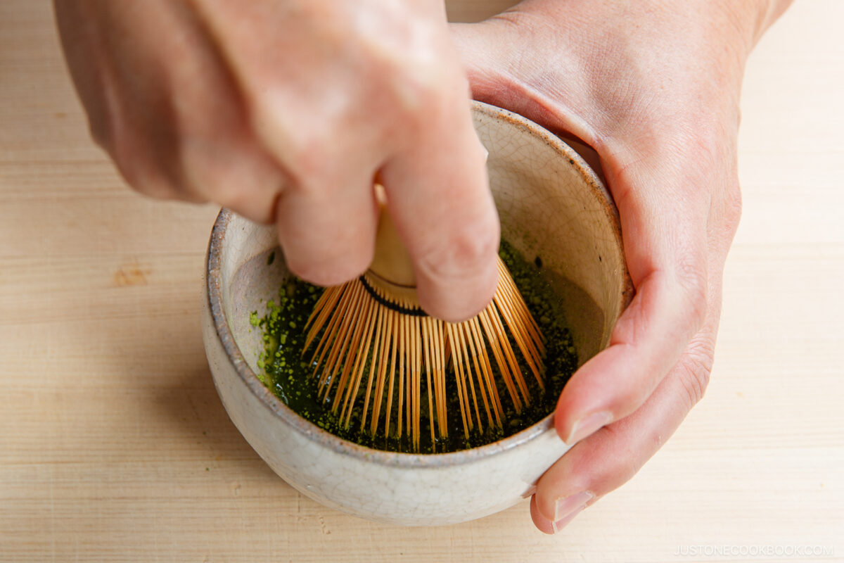 A hand holds a ceramic bowl with green matcha powder, while the other uses a bamboo whisk to mix it on a light wooden surface—perfect for preparing a smooth matcha latte.