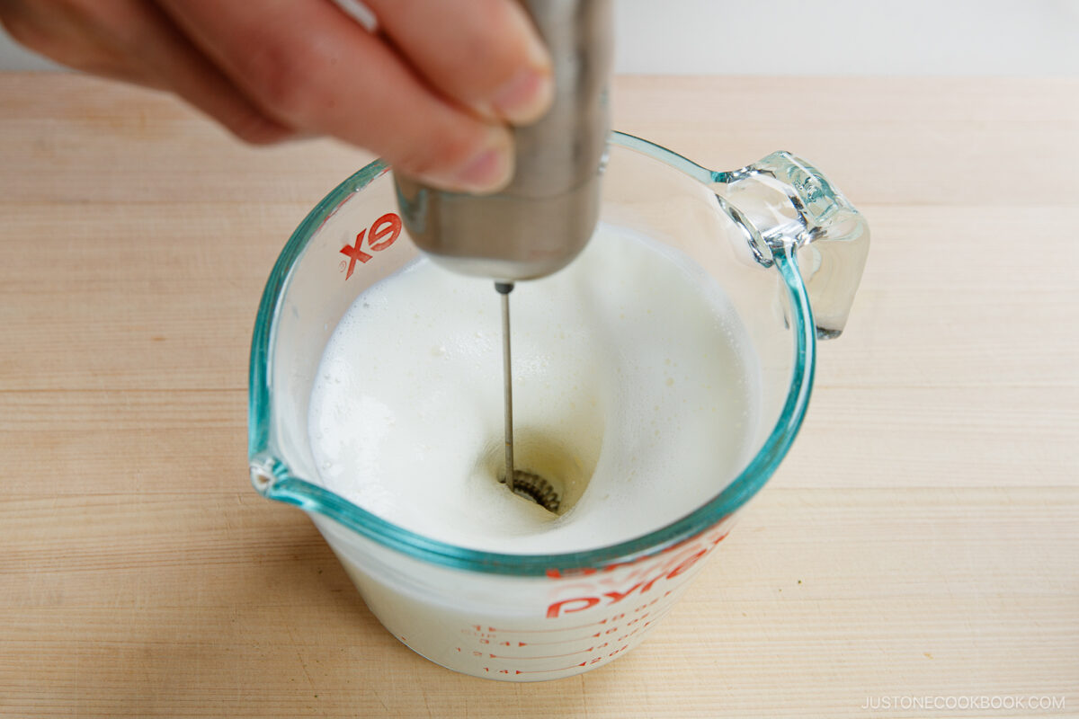A hand uses a handheld milk frother to foam milk for a matcha latte in a glass measuring cup on a wooden surface.