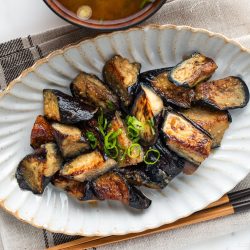 A ceramic plate with grilled eggplant pieces garnished with sliced green onions, evoking the savory flavors of a Miso Eggplant Stir-Fry, is placed next to wooden chopsticks atop a beige cloth. Other dishes and a soup bowl are partially visible around the plate.