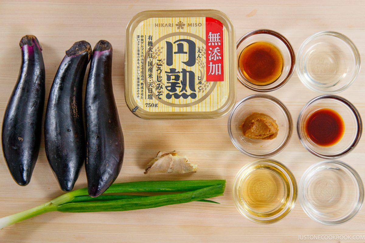 Three Japanese eggplants, a green onion, ginger, miso paste, and small bowls of sake, soy sauce, mirin, and oil are arranged on a wooden surface—everything you need to prepare a flavorful Miso Eggplant Stir-Fry.