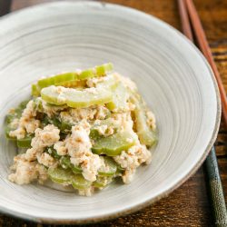 A white bowl contains a dish of sliced green bitter melon mixed with mashed tofu, reminiscent of the freshness found in a Bitter Melon and Tofu Salad. Chopsticks rest on the side of the bowl, which sits on a wooden surface.