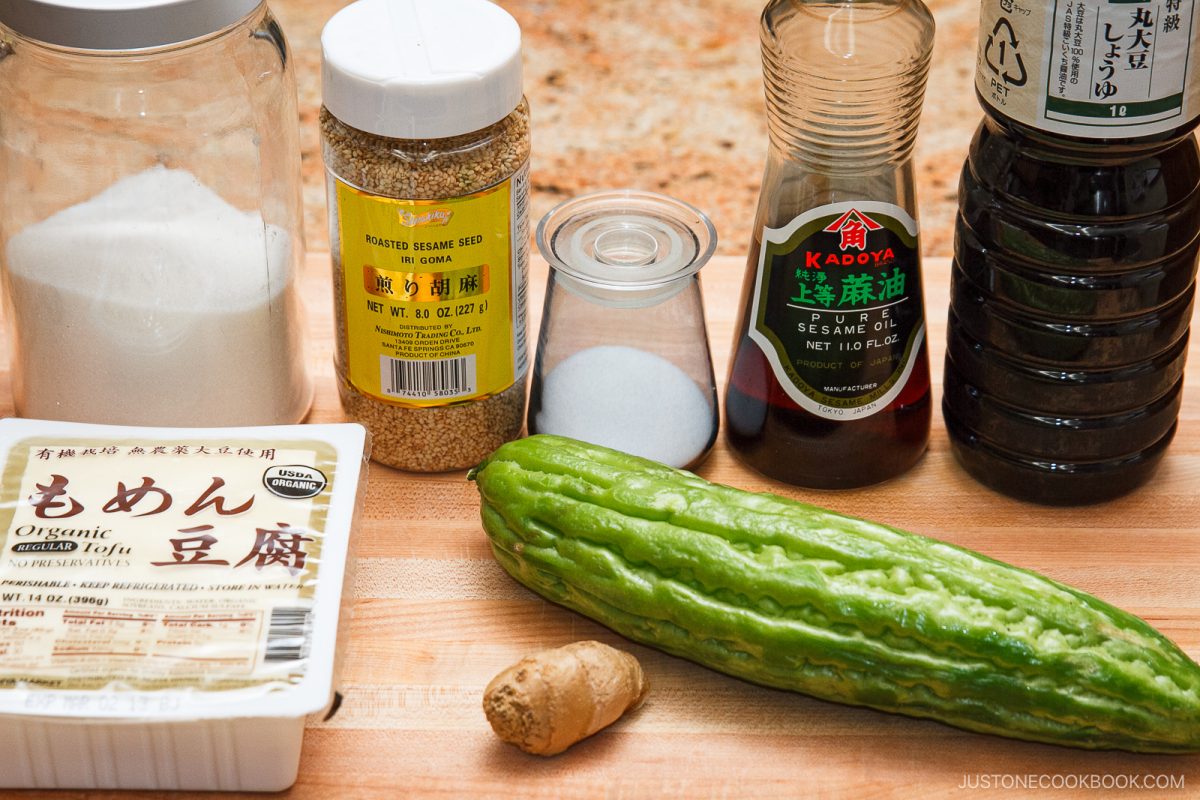 Various ingredients on a wooden countertop, including tofu for a refreshing Bitter Melon and Tofu Salad, a gourd, fresh ginger, a jar of sesame seeds, salt, sugar, soy sauce, and a bottle of sesame oil.