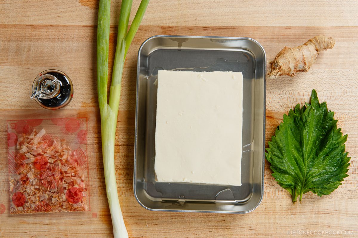 A block of tofu on a tray, with green onions, a piece of ginger, a shiso leaf, a packet of bonito flakes, and a small jar of soy sauce on a wooden surface—classic ingredients for Hiyayakko (Japanese Cold Tofu).