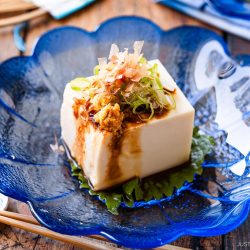 A block of chilled Hiyayakko (Japanese Cold Tofu) topped with green onions, grated ginger, bonito flakes, and soy sauce sits on a shiso leaf in a blue glass dish, with chopsticks and sauce nearby.