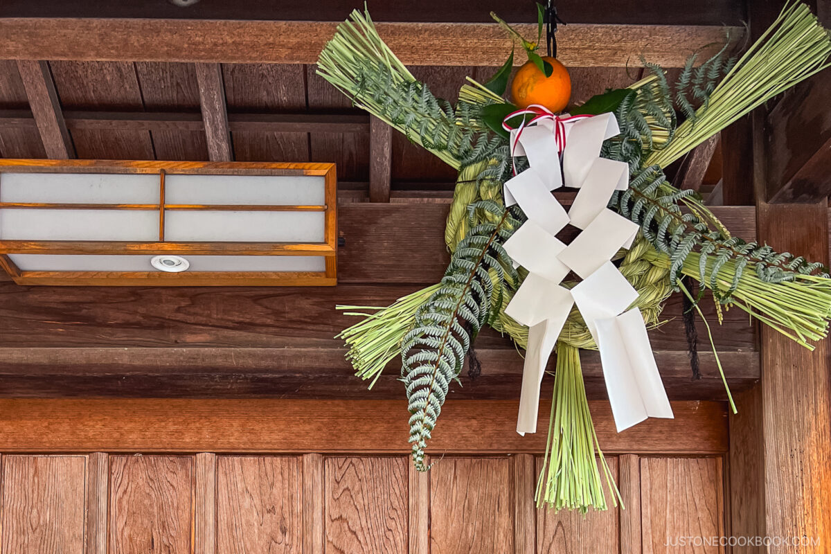 A traditional Japanese shimenawa decoration for Japanese New Year (Oshogatsu), with fern leaves, white paper strips, and an orange, hangs above a wooden door under a wooden roof.