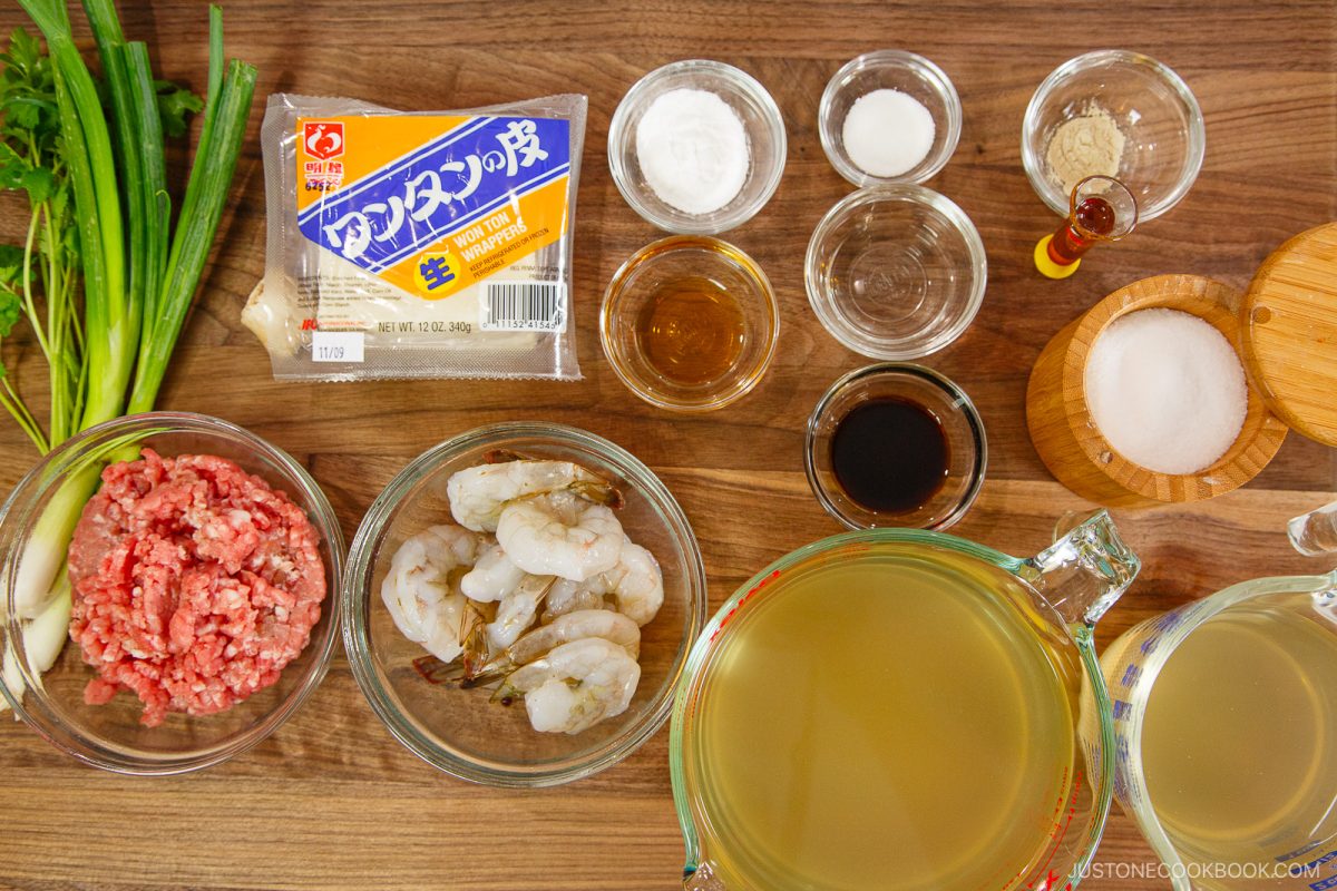 An overhead view of various ingredients for a wonton soup recipe on a wooden table, including ground meat, raw shrimp, green onions, chicken broth, tofu, and small bowls with seasonings and sauces.
