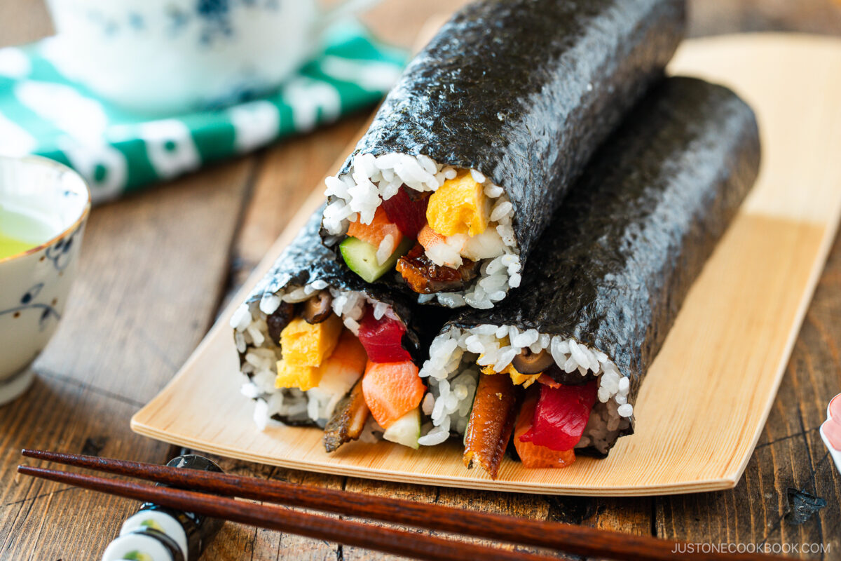 Two uncut sushi rolls—ehomaki (setsubun sushi roll)—filled with rice, vegetables, and seafood are wrapped in nori and arranged on a bamboo plate with chopsticks and a small teacup nearby.