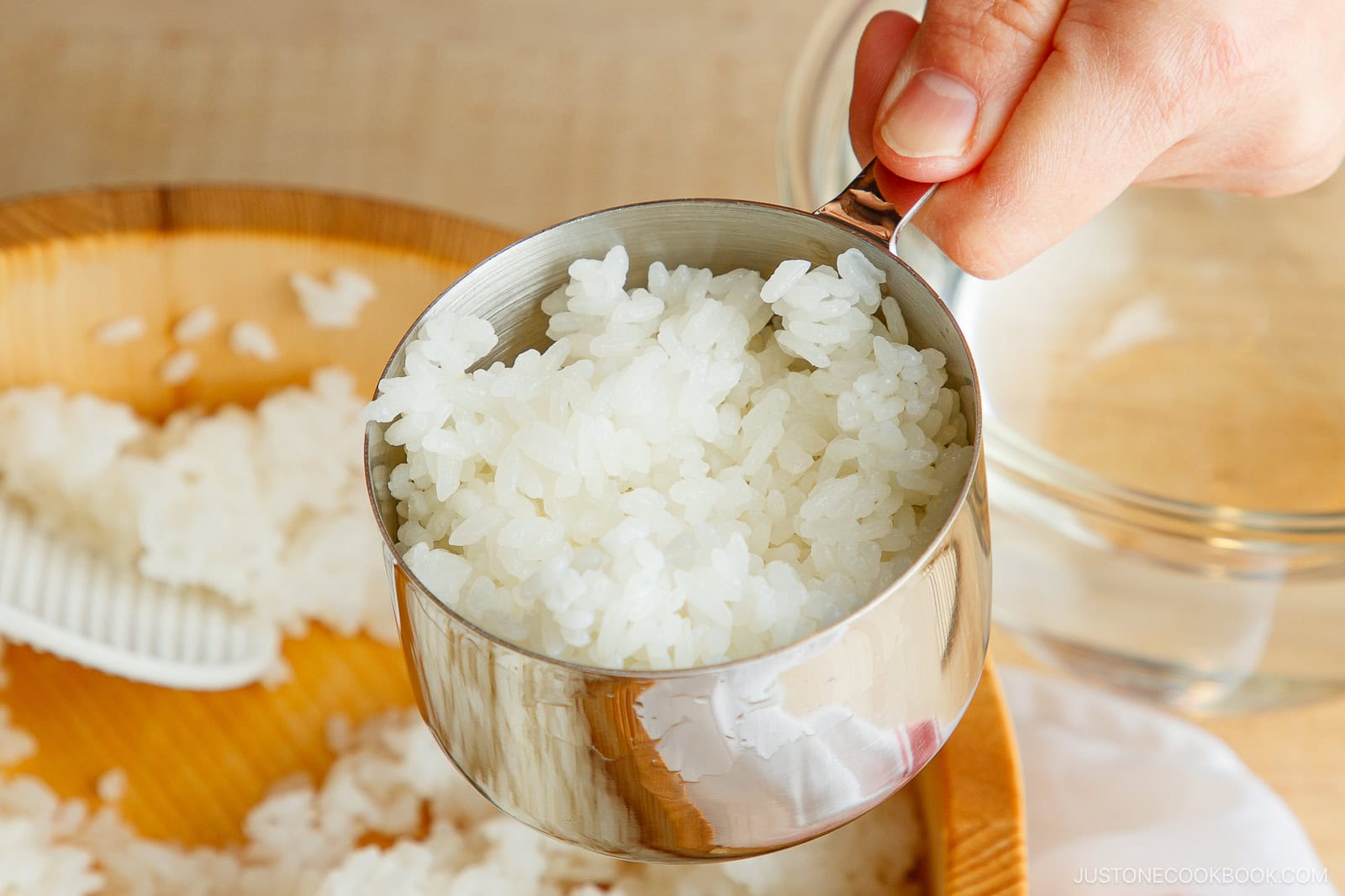 A hand holds a metal measuring cup filled with cooked white rice above a wooden tray, with more rice and a rice paddle—perfect for crafting ehomaki (setsubun sushi roll).