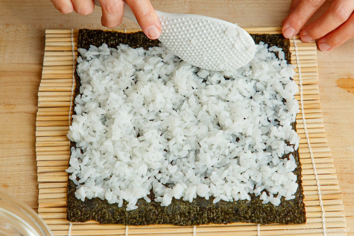 A person spreads white sushi rice over a sheet of nori seaweed on a bamboo sushi rolling mat, using a white rice paddle to prepare ehomaki, the traditional setsubun sushi roll.