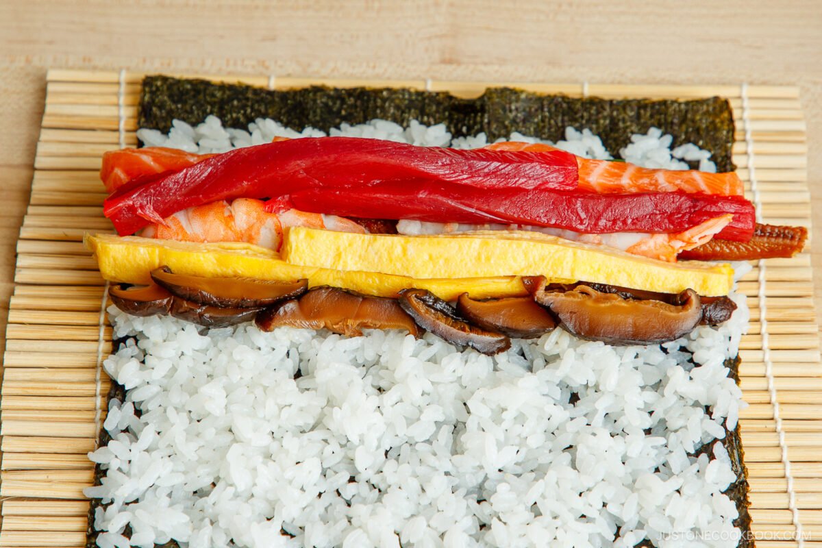 A bamboo sushi mat holds a sheet of nori with sushi rice and layered ingredients—shiitake mushrooms, omelette, shrimp, tuna, salmon, and eel—ready to be rolled into an ehomaki (setsubun sushi roll).