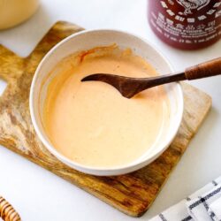 A bowl of creamy orange sauce&mdash;your homemade spicy mayo recipe&mdash;sits with a wooden spoon on a cutting board. Nearby are a halved lime, sriracha bottle, and checkered cloth on a white surface.