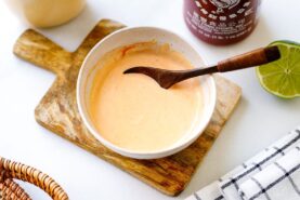 A bowl of creamy orange sauce&mdash;your homemade spicy mayo recipe&mdash;sits with a wooden spoon on a cutting board. Nearby are a halved lime, sriracha bottle, and checkered cloth on a white surface.
