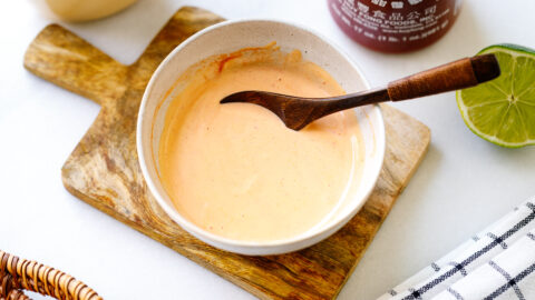 A bowl of creamy orange sauce—your homemade spicy mayo recipe—sits with a wooden spoon on a cutting board. Nearby are a halved lime, sriracha bottle, and checkered cloth on a white surface.