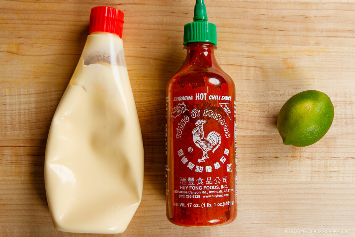 A bottle of mayonnaise, a bottle of Sriracha hot chili sauce, and a whole lime sit on a wooden surface&mdash;perfect ingredients for a homemade spicy mayo recipe.