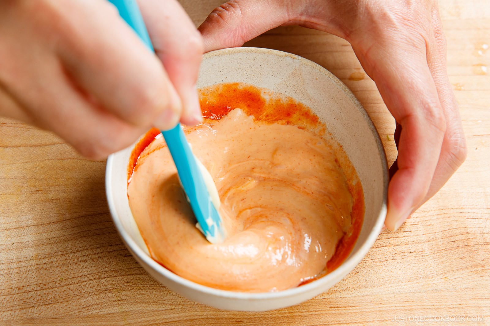 A person uses a blue spatula to mix a creamy, orange-colored homemade spicy mayo recipe in a small white bowl on a wooden surface.