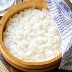 A wooden bowl filled with cooked white sushi rice, perfect for learning how to make sushi rice, sits on a wooden table, partially covered by a blue and white striped cloth with a bamboo sushi mat and ceramic spoon nearby.