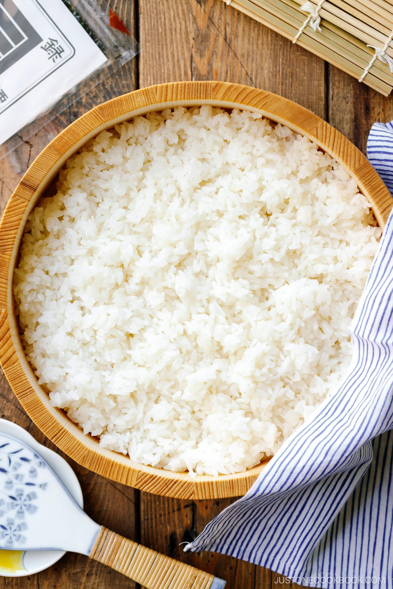 A large wooden bowl filled with cooked white rice sits on a wooden table, partially covered by a blue-and-white striped cloth&mdash;perfect for anyone learning how to make sushi rice. Nearby are a rice paddle and a sushi rolling mat.