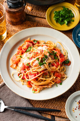 A plate of Tomato Bacon Pasta topped with tomato sauce, fresh chopped tomatoes, grated cheese, and parsley sits on a table set with drinks, a small plate of herbs, and utensils.