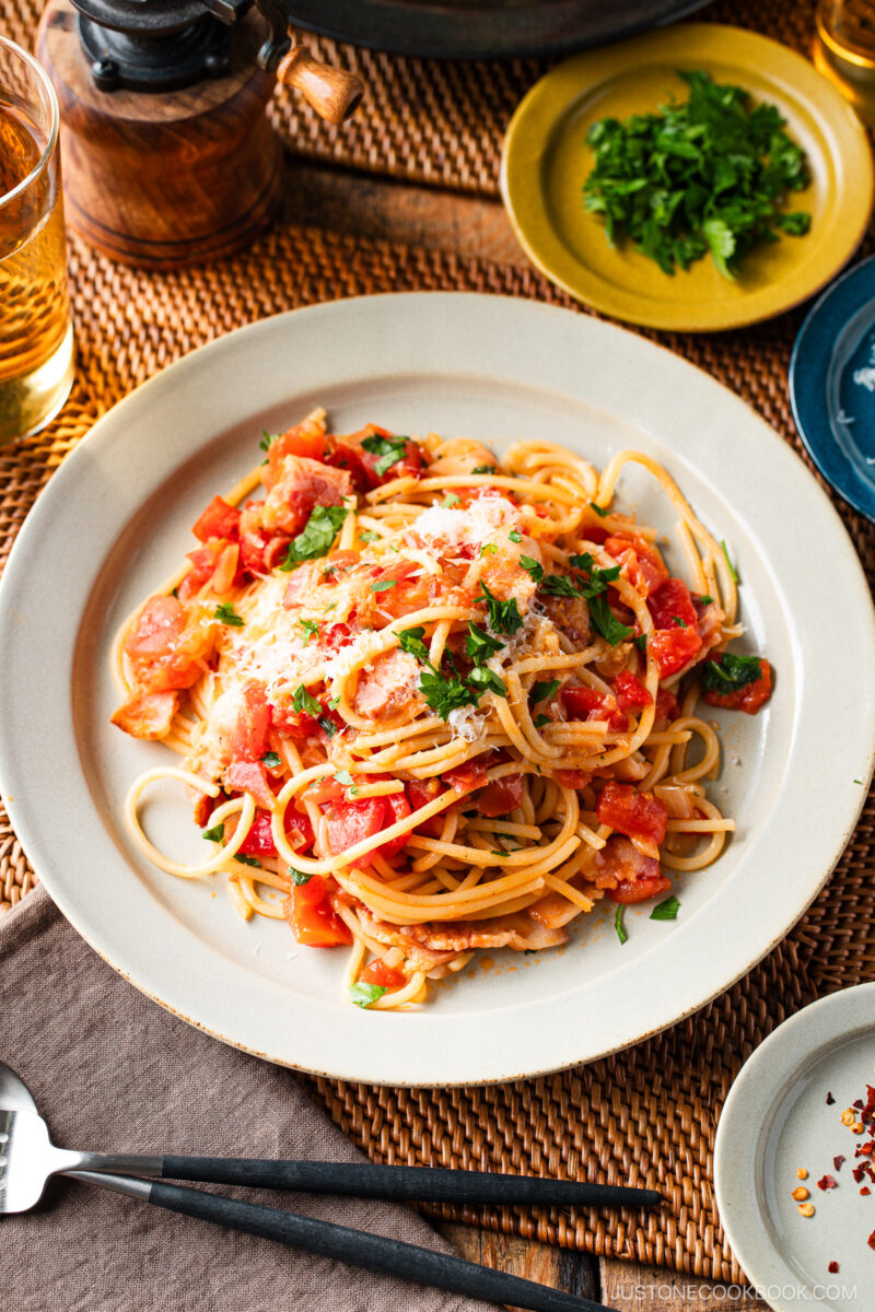 A plate of Tomato Bacon Pasta topped with tomato sauce, fresh chopped tomatoes, grated cheese, and parsley sits on a table set with drinks, a small plate of herbs, and utensils.