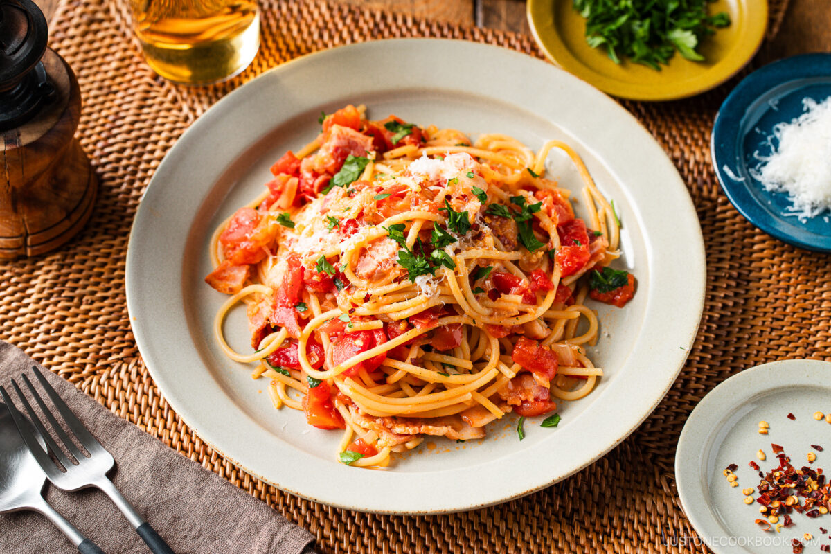 A plate of Tomato Bacon Pasta topped with fresh herbs and grated cheese sits on a woven placemat, with a glass of white wine, extra cheese, herbs, and red pepper flakes on the side. Silverware and a napkin are nearby.