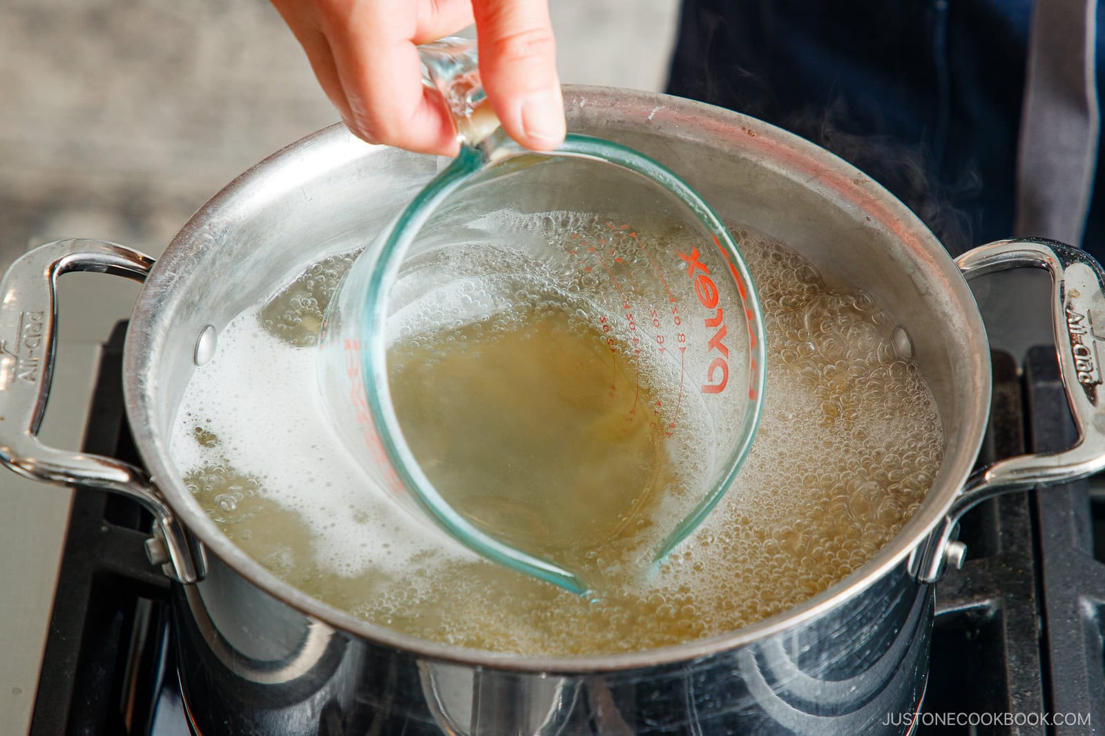 A hand holds a glass measuring cup, scooping hot, cloudy water from a pot of boiling noodles on a stovetop&mdash;perfect for making creamy Tomato Bacon Pasta.