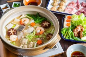 A hot pot of chanko nabe (sumo stew) brims with shrimp, mushrooms, tofu, leafy greens, and vegetables, surrounded by plates of raw meat and dipping sauces&mdash;perfect for communal cooking and sharing at the table.
