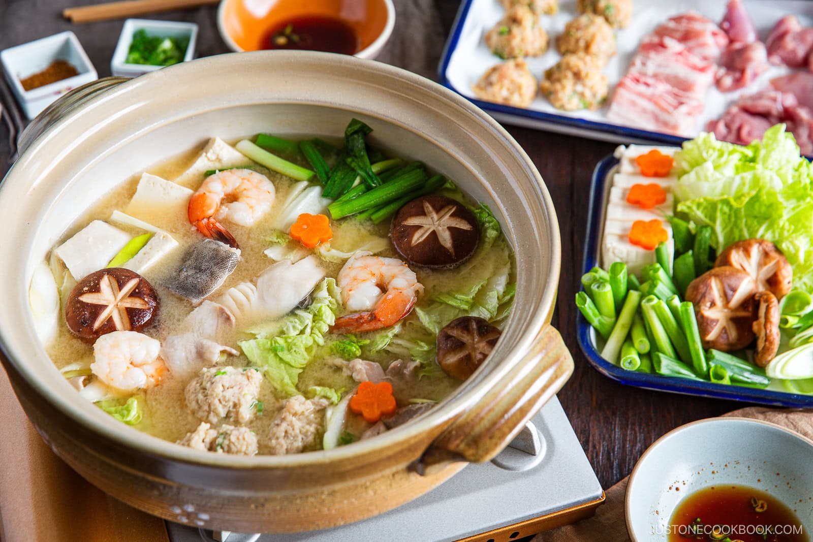 A hot pot of chanko nabe (sumo stew) brims with shrimp, mushrooms, tofu, leafy greens, and vegetables, surrounded by plates of raw meat and dipping sauces—perfect for communal cooking and sharing at the table.