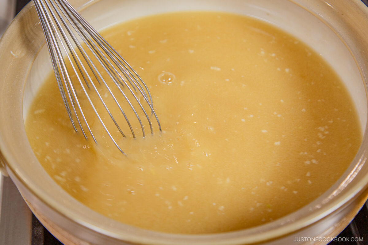 A metal whisk stirring a light brown miso mixture in a beige saucepan on a stovetop, preparing the base for chanko nabe (sumo stew).
