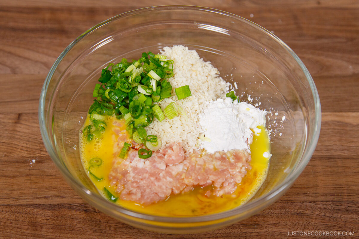 A glass bowl on a wooden surface contains ground chicken, beaten egg, chopped green onions, panko breadcrumbs, and cornstarch—ingredients ready to be mixed for a homemade chanko nabe (sumo stew) recipe.