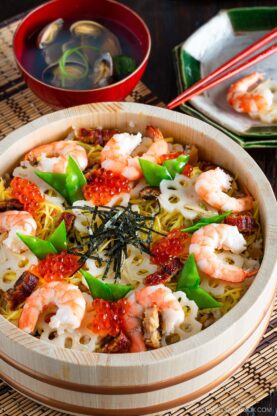A wooden bowl filled with vibrant chirashi sushi, topped with shrimp, ikura (salmon roe), sliced lotus root, green beans, and shredded seaweed. In the background are a bowl of clam soup and a square plate with chopsticks.