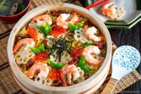 A round wooden dish of chirashi sushi is topped with shrimp, sliced lotus root, snap peas, eel, shredded nori, and bright orange salmon roe. A patterned rice paddle and a plate with chopsticks are placed beside the vibrant chirashi sushi.