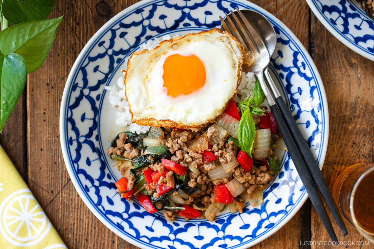 A blue and white patterned bowl filled with gapao rice&mdash;stir-fried ground meat, onions, red bell peppers, basil, and rice&mdash;topped with a sunny-side-up egg. A fork and spoon rest on the side of the bowl.