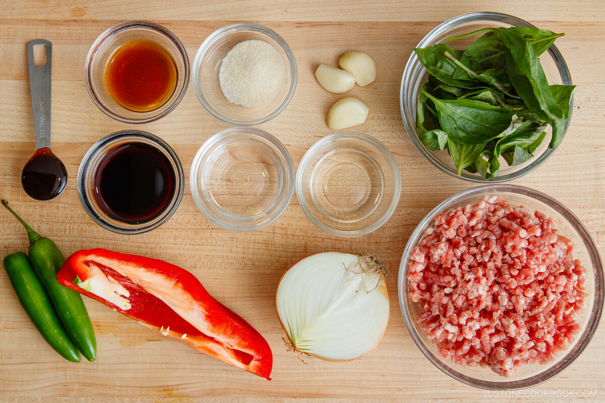 Top-down view of cooking ingredients for gapao rice on a wooden surface, including ground meat, sliced red bell pepper, onion, green chilies, fresh basil, garlic cloves, and small bowls of sauces, sugar, and water.
