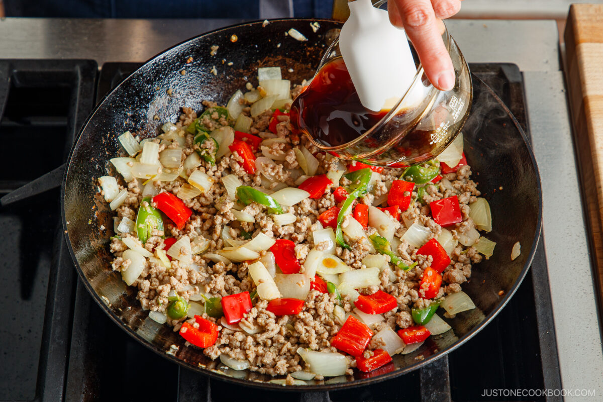 A hand pours dark sauce from a small pitcher into a skillet with cooked ground meat, chopped onions, green bell peppers, and red bell peppers on a stovetop&mdash;perfect flavors for making gapao rice.