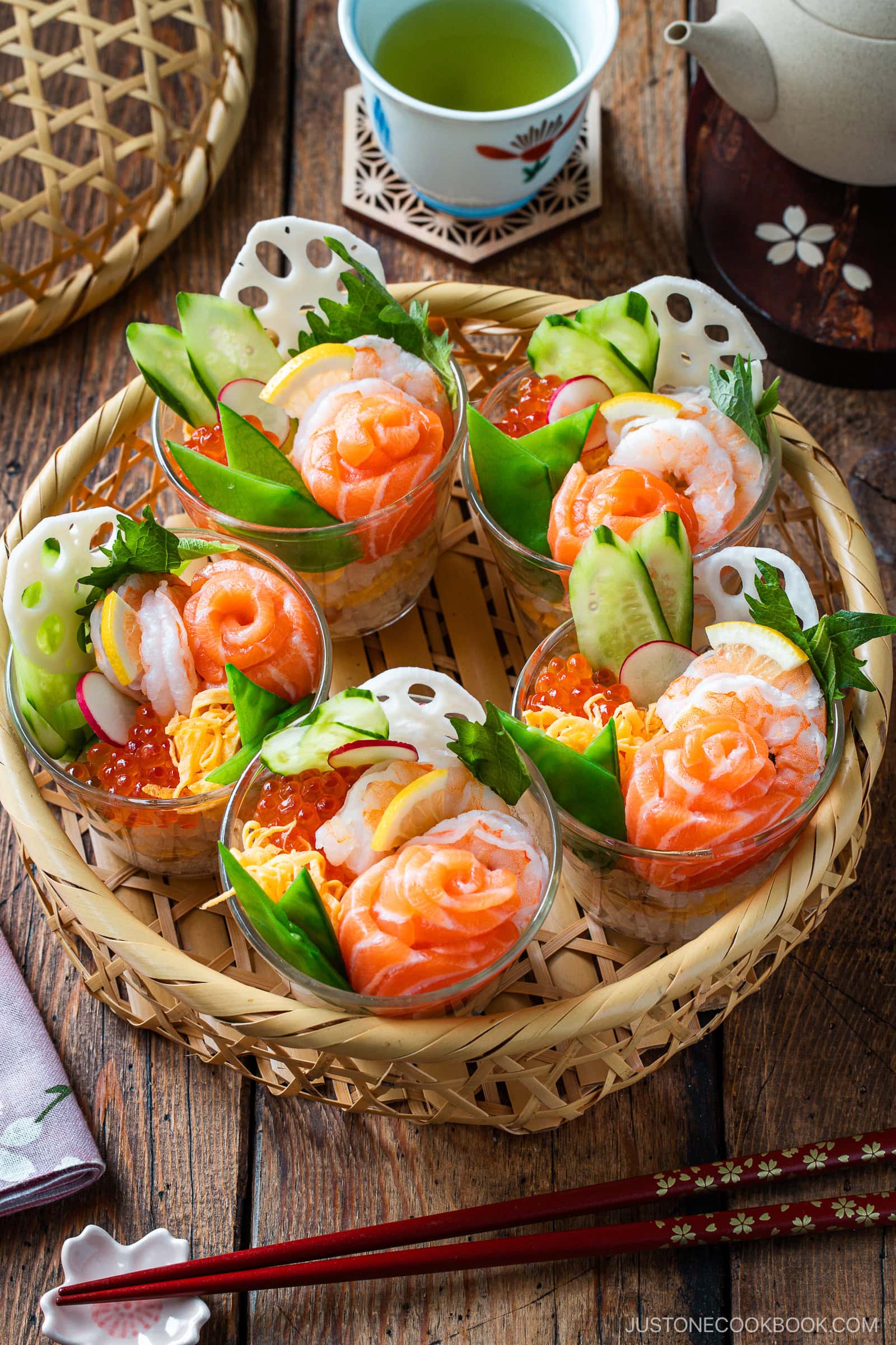 Five clear cups filled with Quick & Easy Chirashi Sushi&mdash;layered with salmon, shrimp, avocado, cucumber, lotus root, radish, and fish roe&mdash;are arranged in a bamboo basket. Green tea and a teapot sit in the background.