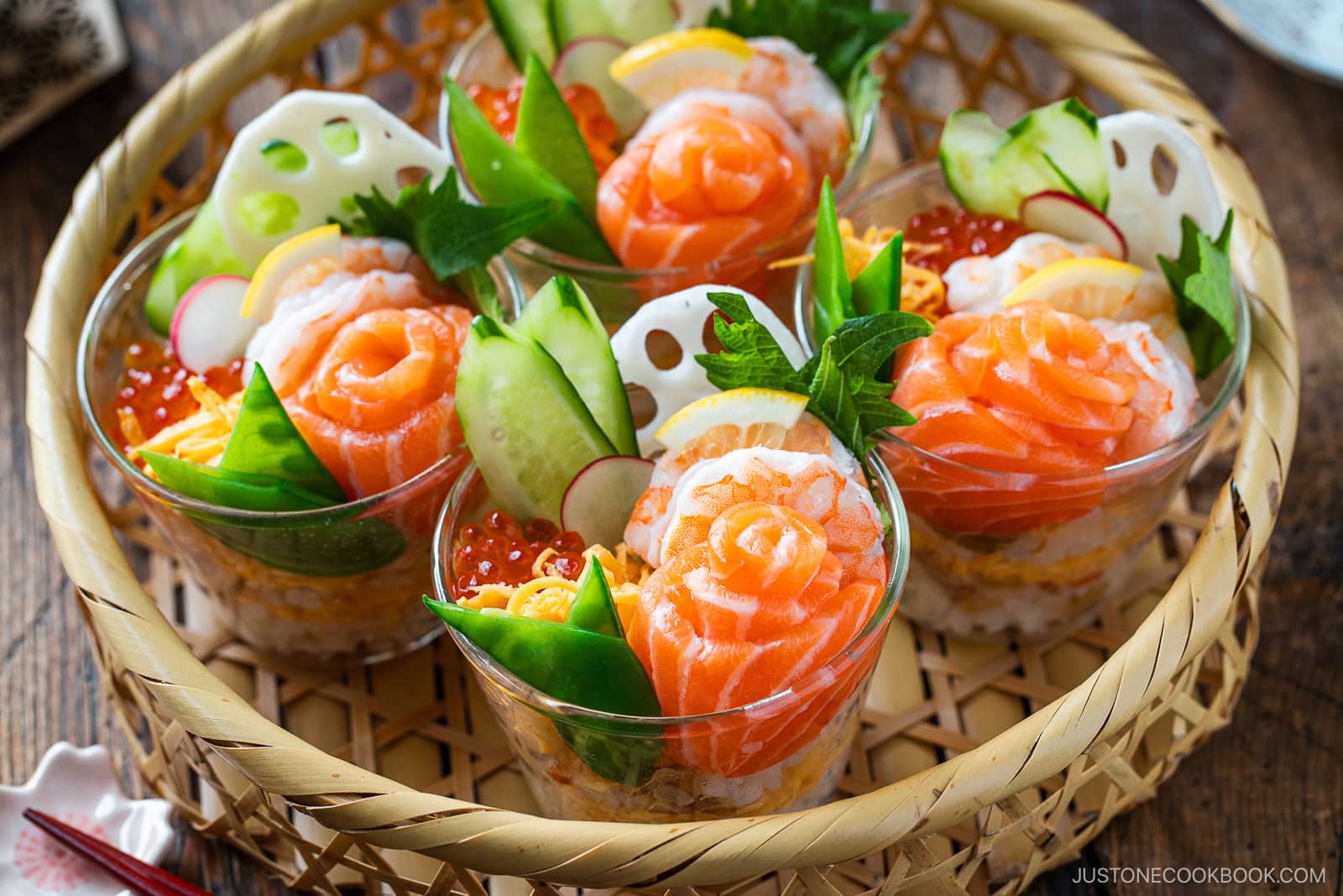 Individual glass cups showcase Quick & Easy Chirashi Sushi, topped with salmon, shrimp, cucumber, radish, snap peas, lotus root, salmon roe, lemon slices, and fresh green garnish&mdash;all artfully arranged in a woven basket.