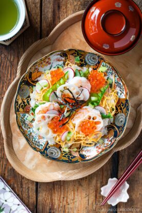 Enjoy a colorful bowl of Quick & Easy Chirashi Sushi topped with shrimp, lotus root, cucumber, radish, shredded egg, and salmon roe, served alongside a red-lidded soup bowl and green tea on a wooden table.