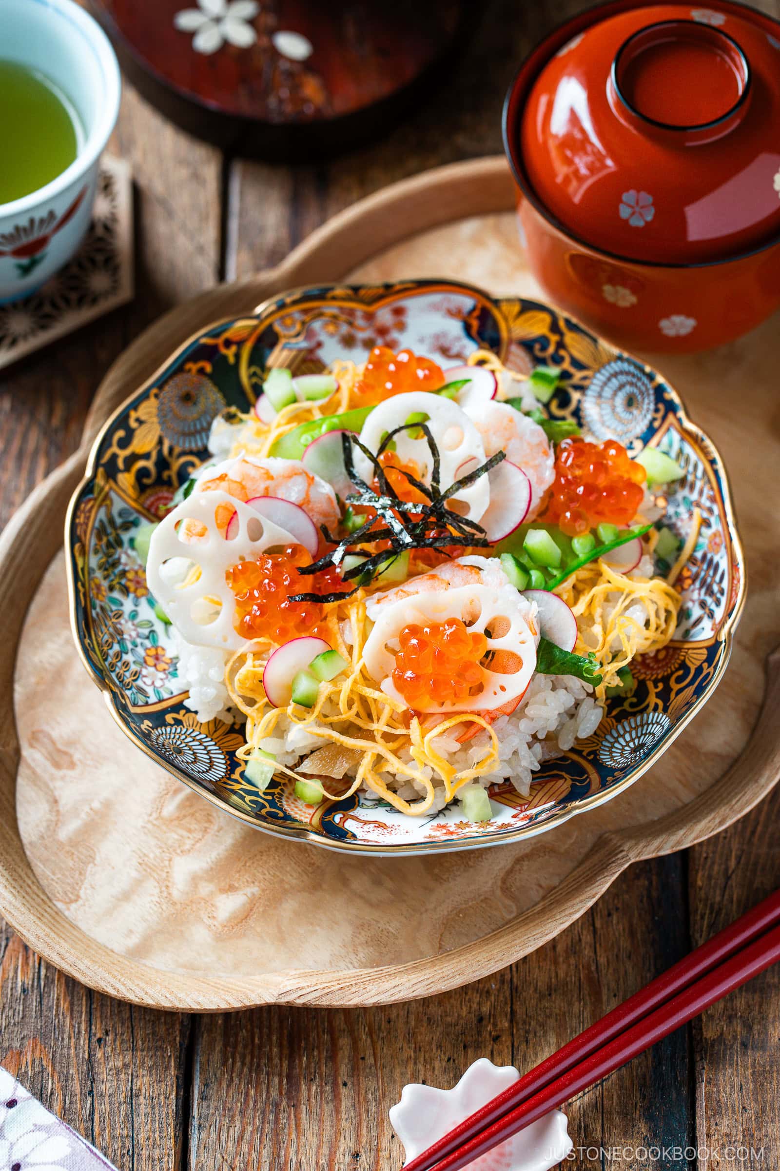 A colorful bowl of Quick & Easy Chirashi Sushi topped with ikura (salmon roe), sliced lotus root, shredded egg, cucumber, and seaweed sits on a wooden tray next to a cup of green tea and a miso soup bowl.