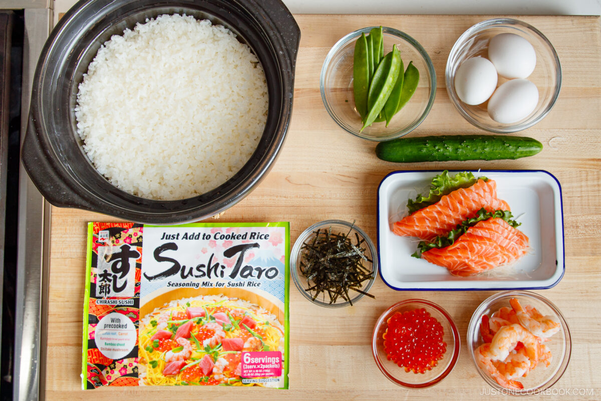 A wooden surface holds cooked rice in a pot, a cucumber, snap peas, eggs, raw salmon slices, shrimp, salmon roe, shredded seaweed, and a box of Sushi Taro seasoning mix&mdash;everything you need for Quick & Easy Chirashi Sushi.
