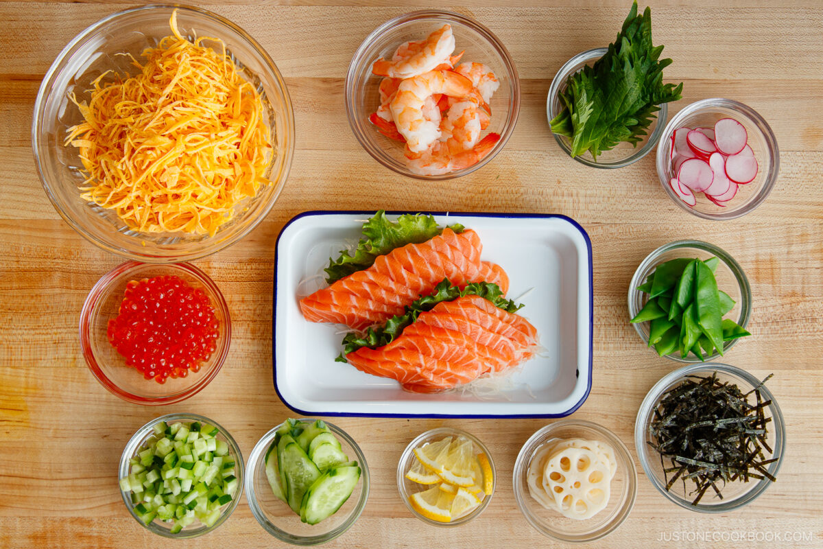 An overhead view of assorted Quick & Easy Chirashi Sushi toppings in bowls, featuring salmon slices, shrimp, shredded egg, shiso leaves, radishes, cucumber, snap peas, lemon, lotus root, seaweed strips, salmon roe, and sliced cucumber.