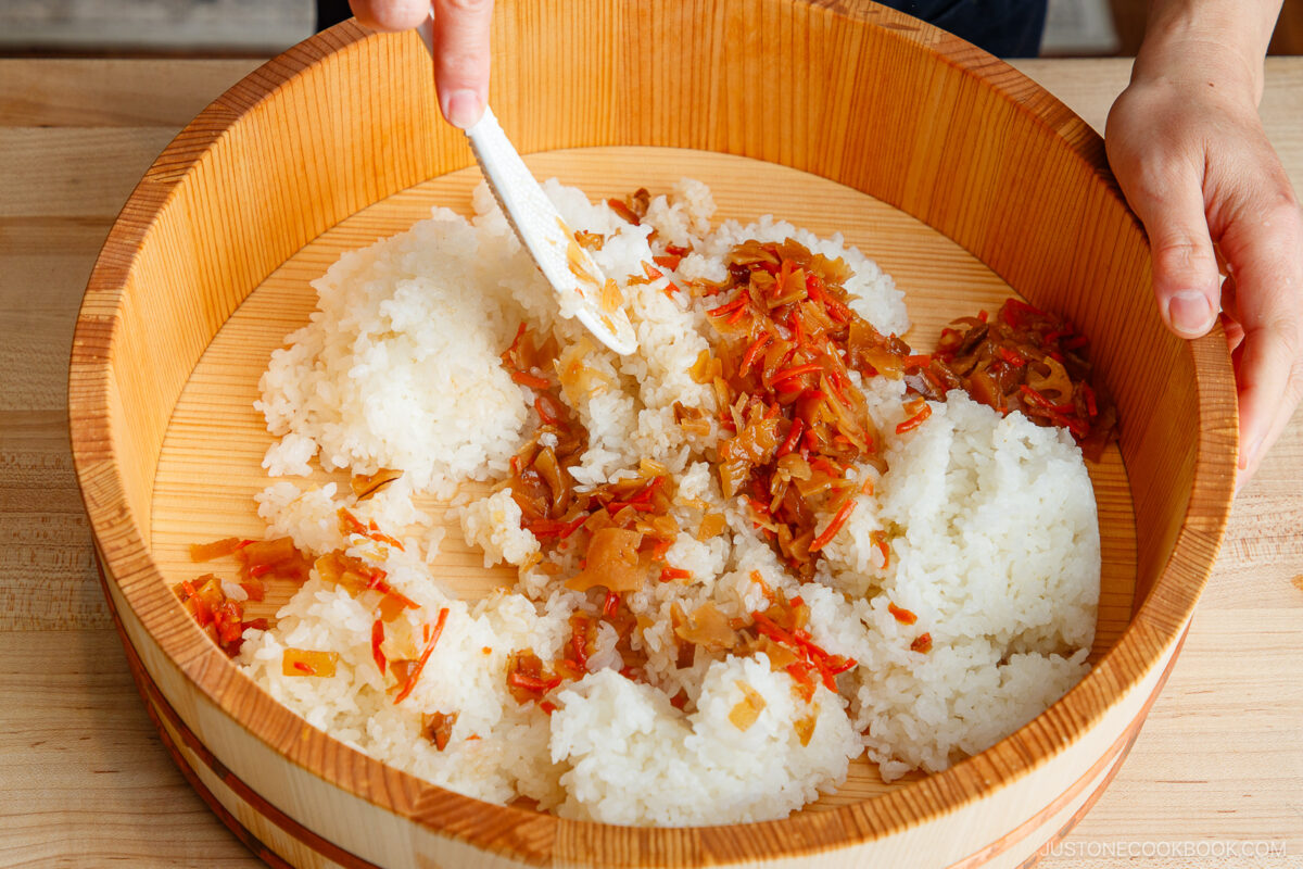 A person prepares Quick & Easy Chirashi Sushi by mixing cooked white rice with chopped vegetables and seasoning in a large wooden bowl using a spatula. The ingredients are spread evenly across the bowl&rsquo;s surface.