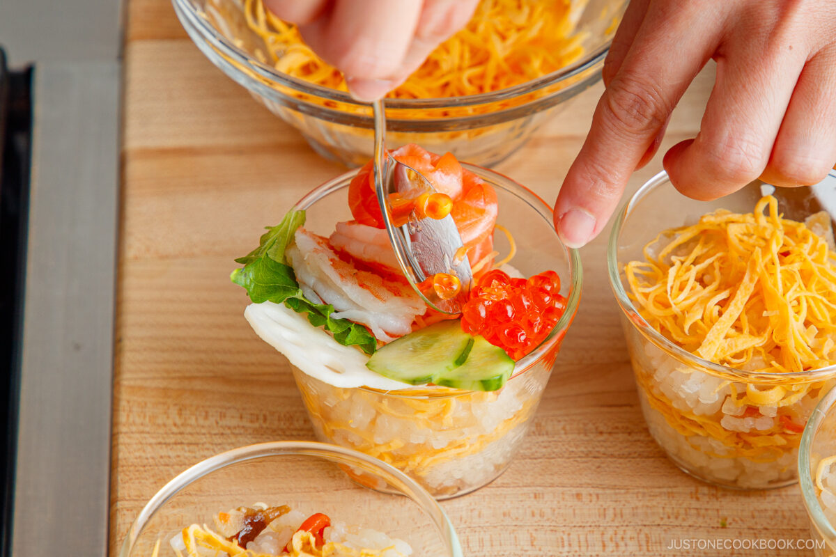A hand uses a fork to arrange sliced fish, cucumber, and salmon roe on top of a small glass filled with rice and shredded egg&mdash;creating Quick & Easy Chirashi Sushi&mdash;with similar cups in the background on a wooden surface.