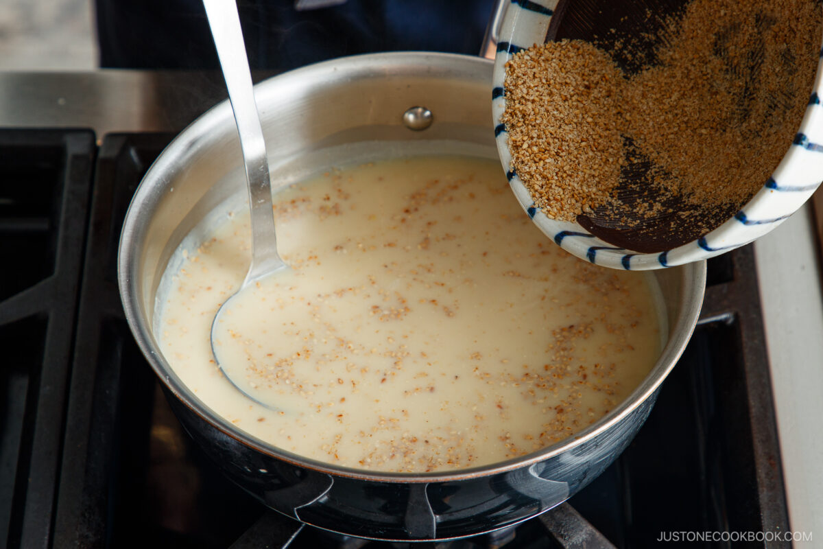 A person pours ground sesame seeds from a bowl into a saucepan of creamy liquid on a stovetop, using a ladle to stir the mixture for tan tan udon.