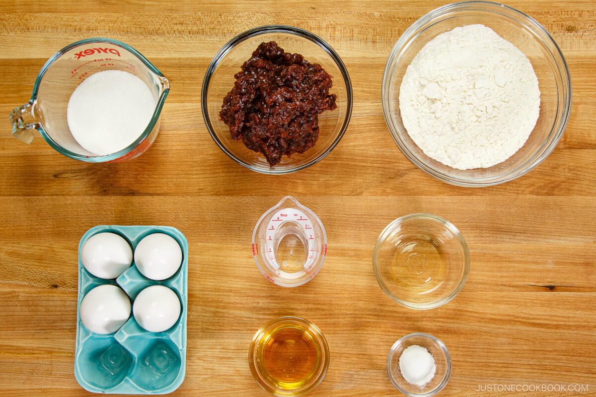 Baking ingredients arranged on a wooden surface for dorayaki (Japanese red bean pancake): sugar, flour, eggs, chocolate mixture, oil, water, vanilla extract, and salt&mdash;each neatly placed in separate bowls or measuring cups in an overhead layout.