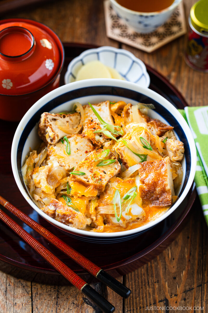 A bowl of Japanese Simmered Tofu and Egg Rice Bowl with fried tofu, egg, onions, and scallions sits on a tray next to red chopsticks, pickled ginger, a lidded bowl, and a cup of tea on a wooden table.