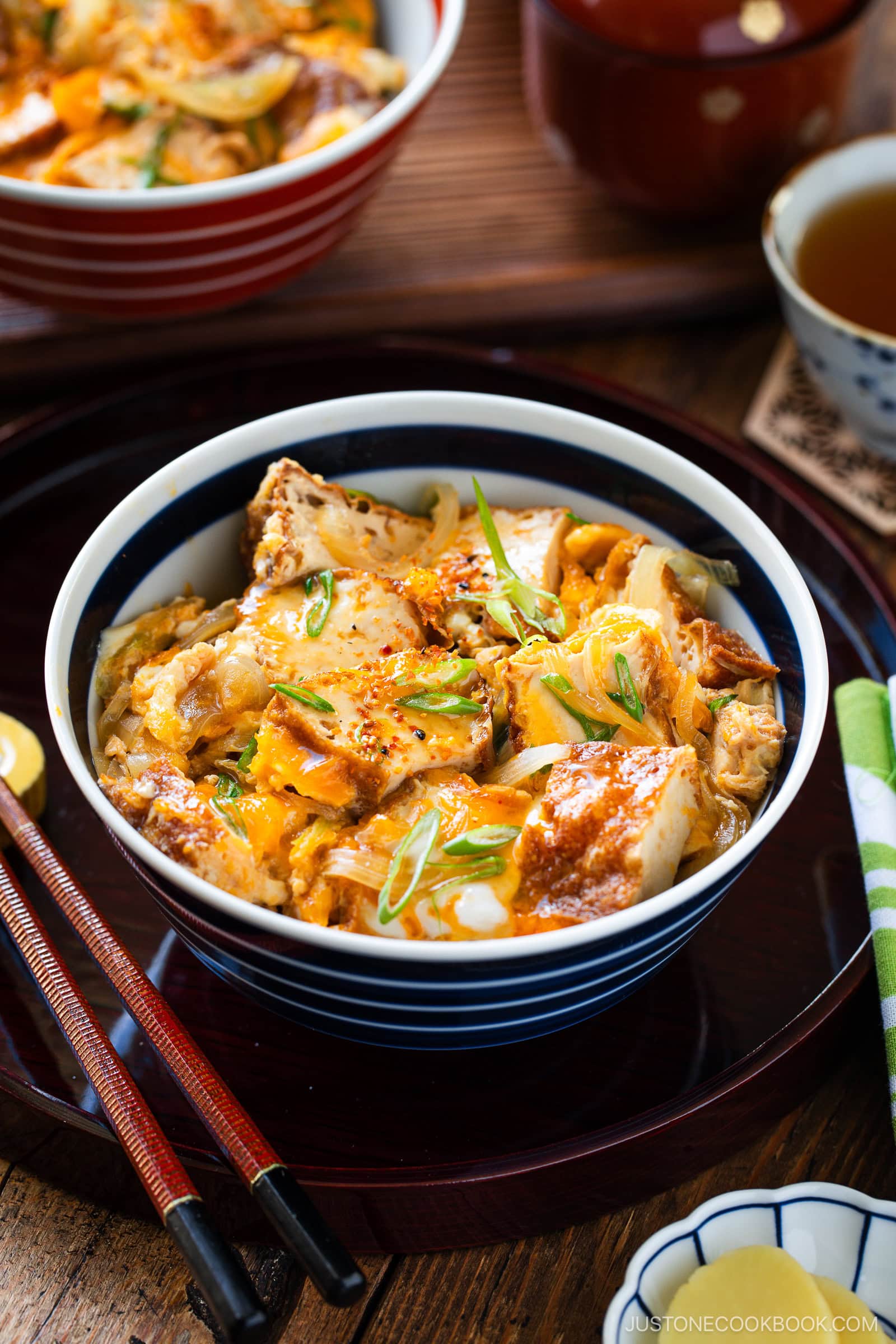 A bowl of Simmered Tofu and Egg Rice Bowl, similar to oyakodon, features seasoned fried tofu, eggs, and onions over steamed rice, garnished with sliced green onions. Chopsticks rest on the tray beside tea and soup in the background.