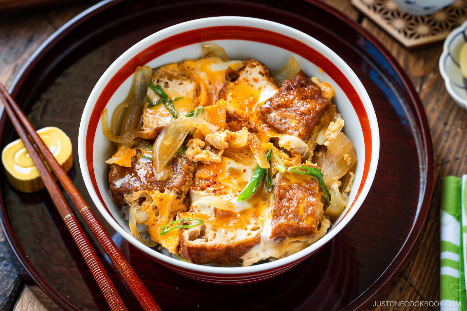 A bowl of Japanese simmered tofu and egg rice bowl with tender onions, and scallions, garnished with spices, set on a tray with chopsticks and a flavorful side dish.
